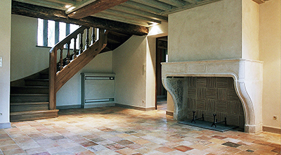Aménagement d’un salon avec cheminée en pierre, carrelage en terre cuite ancienne et escalier reconditionné.